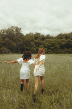 two women in white dresses are walking through the tall grass with trees in the background