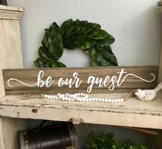 a wooden sign that says be our guest on top of a shelf next to a potted plant