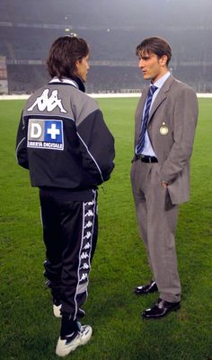 two men in suits standing on a soccer field talking to each other and looking at the camera