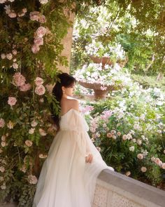 a woman in a white dress leaning against a wall with flowers on it and greenery behind her