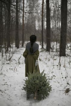 a woman walking through the woods carrying a small christmas tree in her hand and wearing a cloak