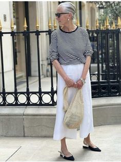 an older woman is standing on the sidewalk with her hand in her pocket and holding a straw bag