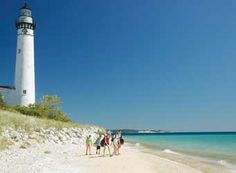 several people are walking on the beach near a light house in the ocean with clear blue skies