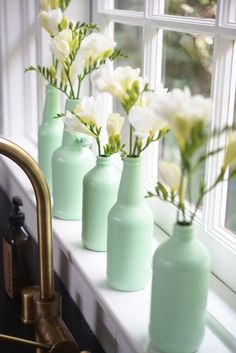 four green vases with white flowers in them sitting on a window sill next to a faucet