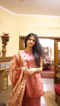 a woman standing in a room with long black hair wearing a pink dress and shawl