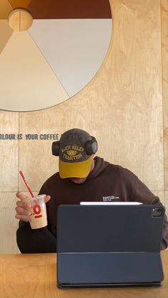 a man sitting in front of a laptop computer wearing a hat and holding a drink