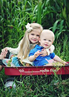 two young children sitting in a red wagon with grass and plants behind them, both smiling at the camera