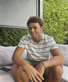 a young man sitting on top of a bed next to a green bushy wall