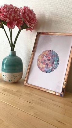 pink flowers are in a vase next to a framed photograph on a wooden table with a white wall behind it