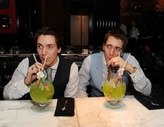 two men sitting at a table with drinks in front of their faces and holding straws up to their mouths