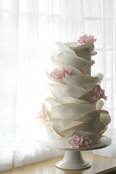 a three tiered white cake with pink flowers on the top and bottom, sitting on a table in front of a window