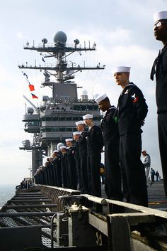 a group of sailors standing on the deck of a ship looking at an aircraft carrier