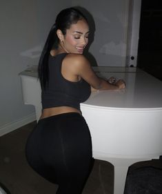 a woman in tight black pants leaning on a white piano