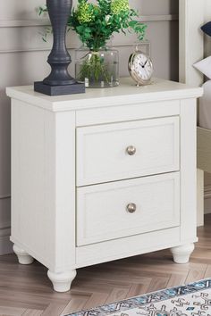 a white night stand with two drawers and a clock on the top, next to a plant