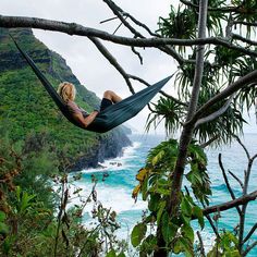 a woman laying in a hammock on top of a tree next to the ocean