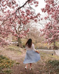 a woman in a blue dress walking down a path with pink flowers on the trees