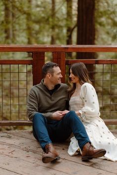 a man and woman sitting on a wooden deck in front of trees, smiling at each other