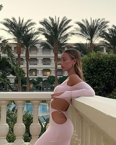 a woman in a pink dress leaning on a balcony railing with palm trees behind her