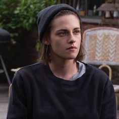 a woman sitting on the ground wearing a black sweater and a beanie with her hands in her pockets