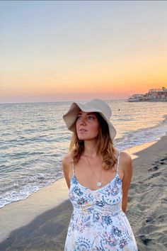 a woman standing on top of a sandy beach next to the ocean wearing a hat