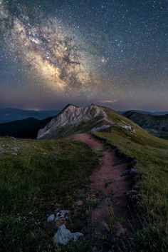 the night sky is filled with stars above a grassy field and mountain side, as well as a trail leading up to it