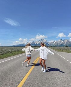 two girls are walking down the road in front of mountains and sky with their arms outstretched