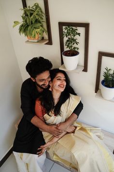 a man and woman sitting next to each other on top of a wooden bench in front of potted plants