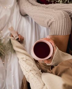 A brand photographer captures the serene moment of a person holding a pink mug with a warm beverage, wearing cozy knit socks, and sitting on a bed with a knitted blanket. Knit Stockings, Cozy Autumn