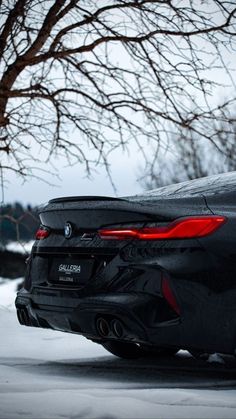 the rear end of a black sports car parked in front of a tree on a snowy day