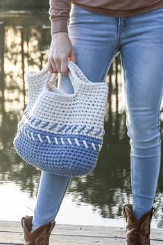 a woman is holding a crocheted purse by the water's edge while wearing jeans and boots