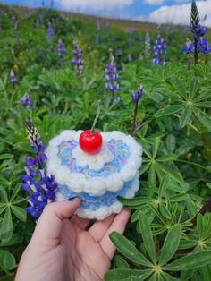 a hand is holding a miniature cake with a cherry on it in the middle of flowers