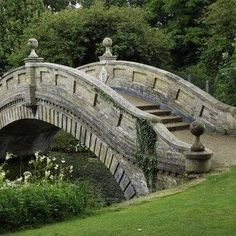 an old stone bridge in the middle of a park with flowers and trees around it