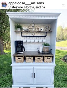 a white hutch with coffee maker and baskets on it in the grass next to a tree