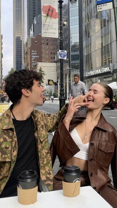 a man and woman sitting at a table with coffee cups in front of them on the street