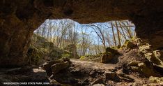 a cave entrance in the woods with rocks and trees on either side, looking out into the distance