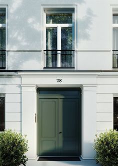 a white building with two green doors and some bushes on the sidewalk in front of it