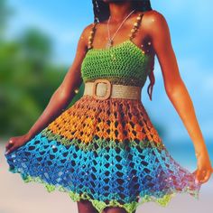 a woman wearing a colorful crochet dress with a belt around her waist, standing in front of a white wall