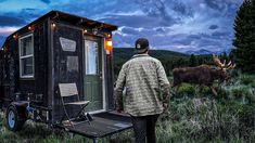 a man standing next to a trailer in the grass with an elk behind him at night