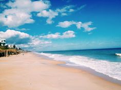 a sandy beach next to the ocean under a blue sky with white clouds and houses