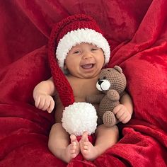 a baby wearing a santa hat and holding a teddy bear sitting on a red blanket