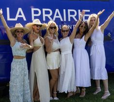 four women in white dresses and hats posing for the camera with their arms up while standing next to a blue bus