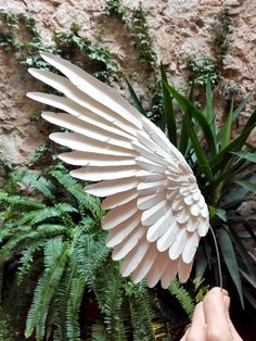 a hand is holding a white paper angel wing in front of some plants and rocks
