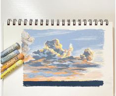 some crayons are laying on top of a drawing book with clouds in the background