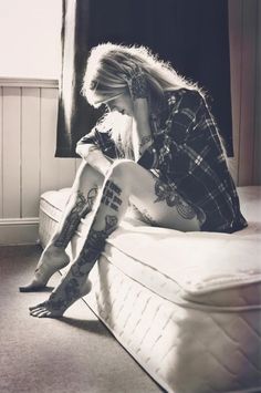 a woman sitting on top of a bed with tattoos on her arms and legs, next to a window