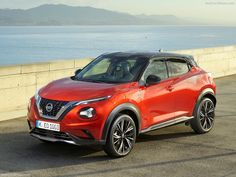 a red nissan juke is parked on the side of the road by the water