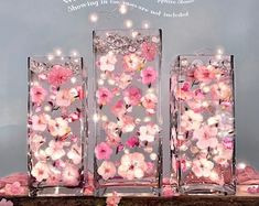 three clear vases filled with pink flowers on top of a wooden table covered in fairy lights