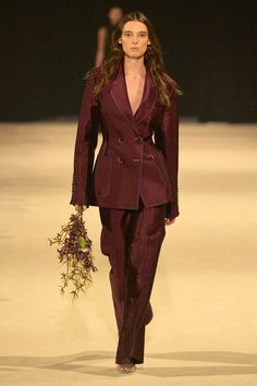 a woman walking down a runway wearing a suit and holding a bouquet of flowers in her hand