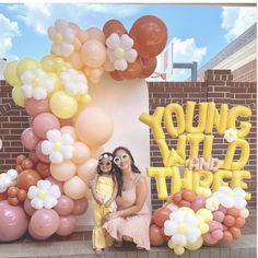 two women sitting in front of a large balloon arch with the words young wild things on it