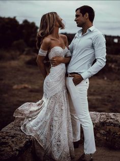 a man and woman standing next to each other on top of a rocky hill with the ocean in the background