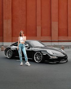 a woman standing next to a black sports car in front of a tall building with red walls